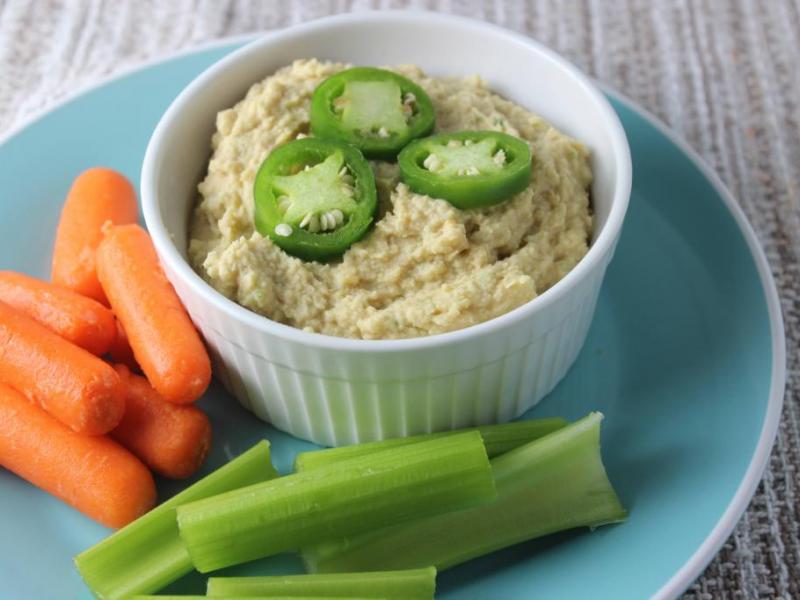 Party Hummus topped with fresh jalapeno slices in a white dish served with carrot and celery sticks